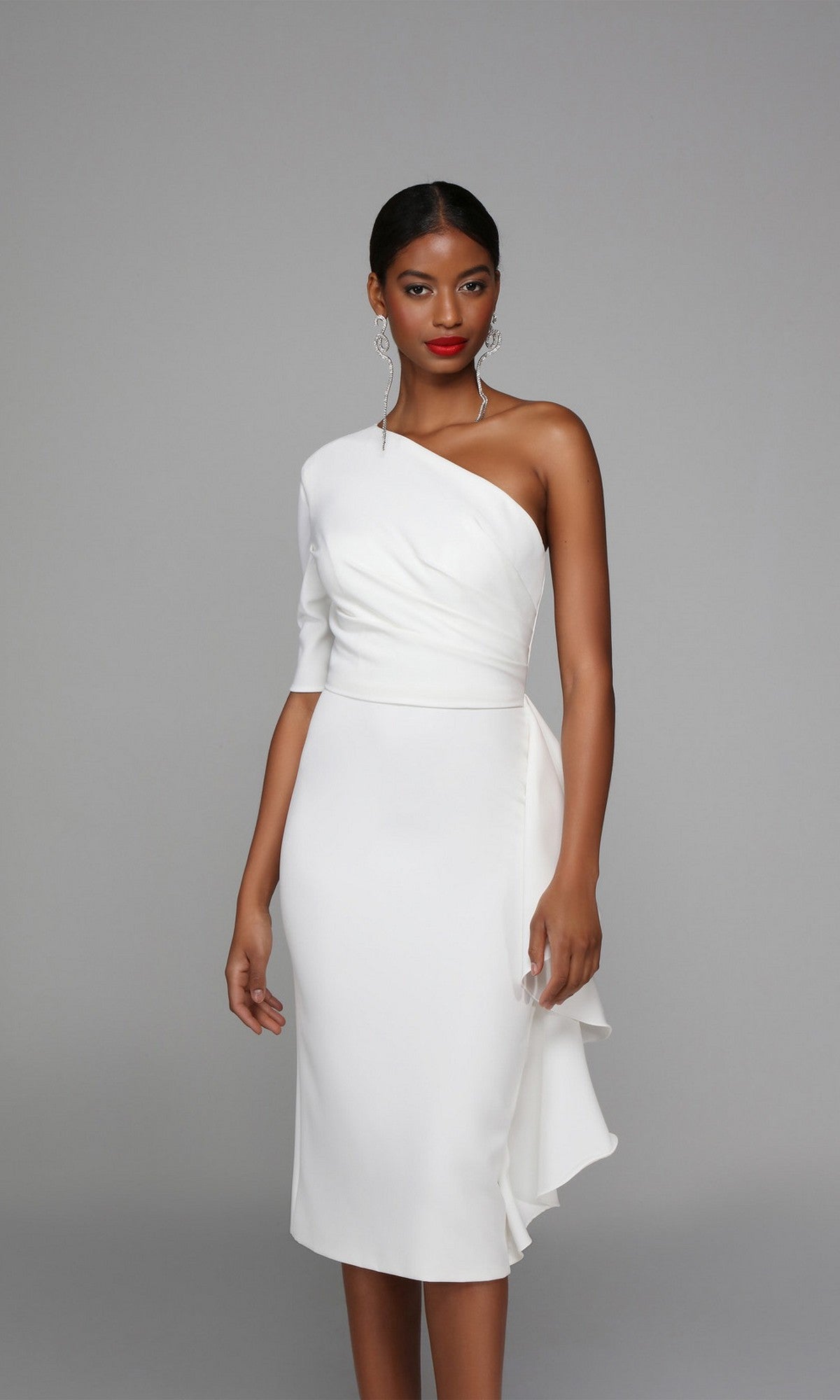 A woman poses against a gray background wearing the elegant Alyce 70006 knee-length semi-formal dress, featuring a white one-shoulder design with ruffled detail, paired with drop earrings.